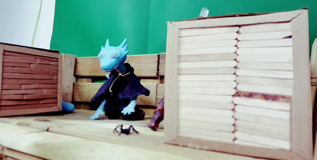 A photo showing the artist’s puppet of her original character, Liz, on the set of her final animation for university. The model is made from polymer clay, plastercine and wire, with hand-sewn clothes. The set is hand-made using cardboard and wood to create