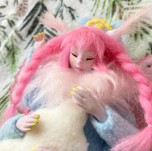 A closeup photo of a sculpture of another artist’s (Chaoillustrations) original character, Mallow. She’s a pink skinned anthropomorphic moth, with four arms and twin braids. She is wearing a blue fleece onesie and holding a fleece cloud pillow while sleepi